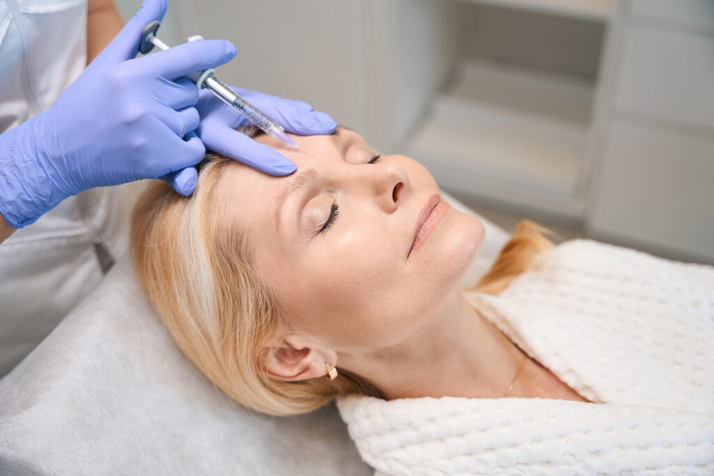 Close up of cosmetologist hands performing rejuvenating injection on mature lady forehead in beauty salon
