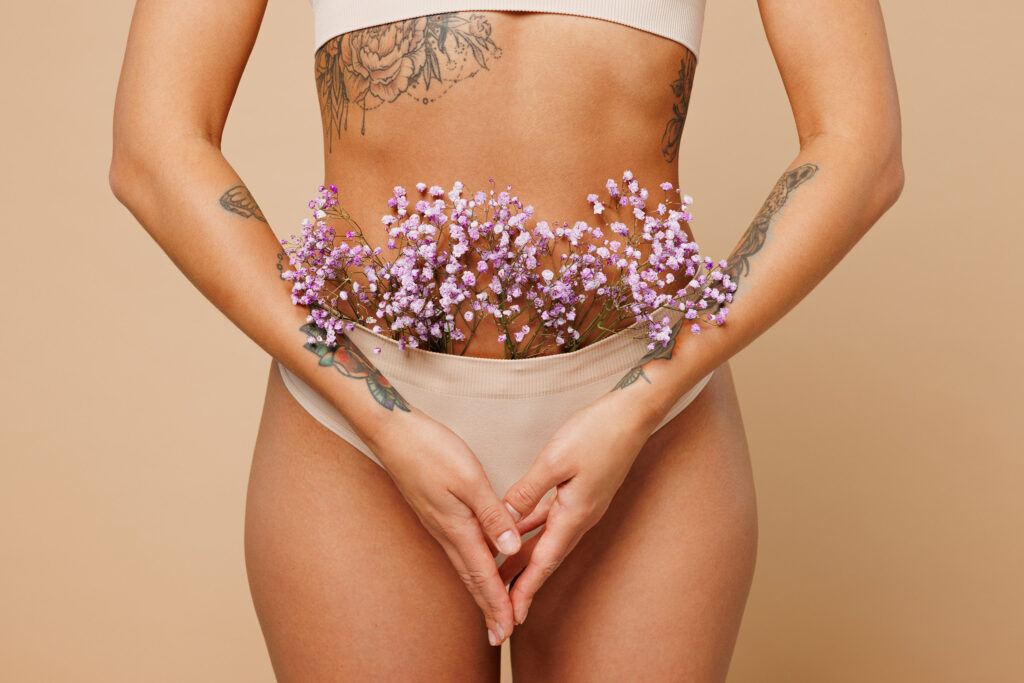 Close up cropped young lady woman with slim body perfect skin wearing nude top bra lingerie standing with flower in panties isolated on plain pastel light beige background.